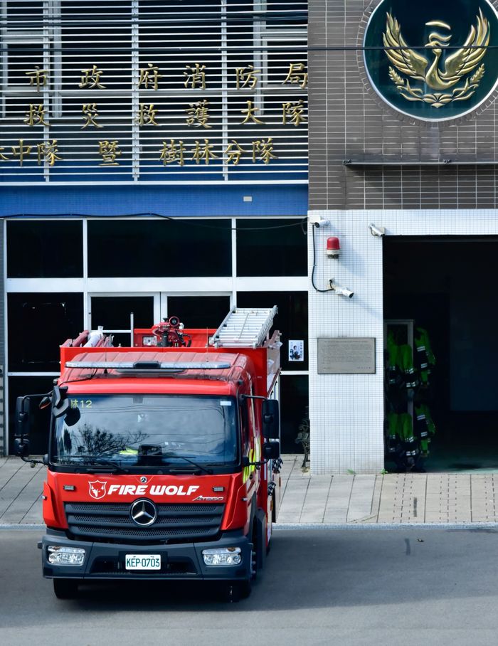 gallery-5 Red firetruck parked outside a fire station with a distinctive emblem on the building.